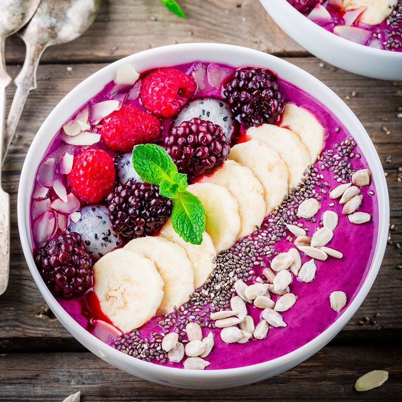 Schüssel mit Smoothie, Himbeeren, Brombeeren, Bananenscheiben, Samen und Mandeln. Auf Holz.