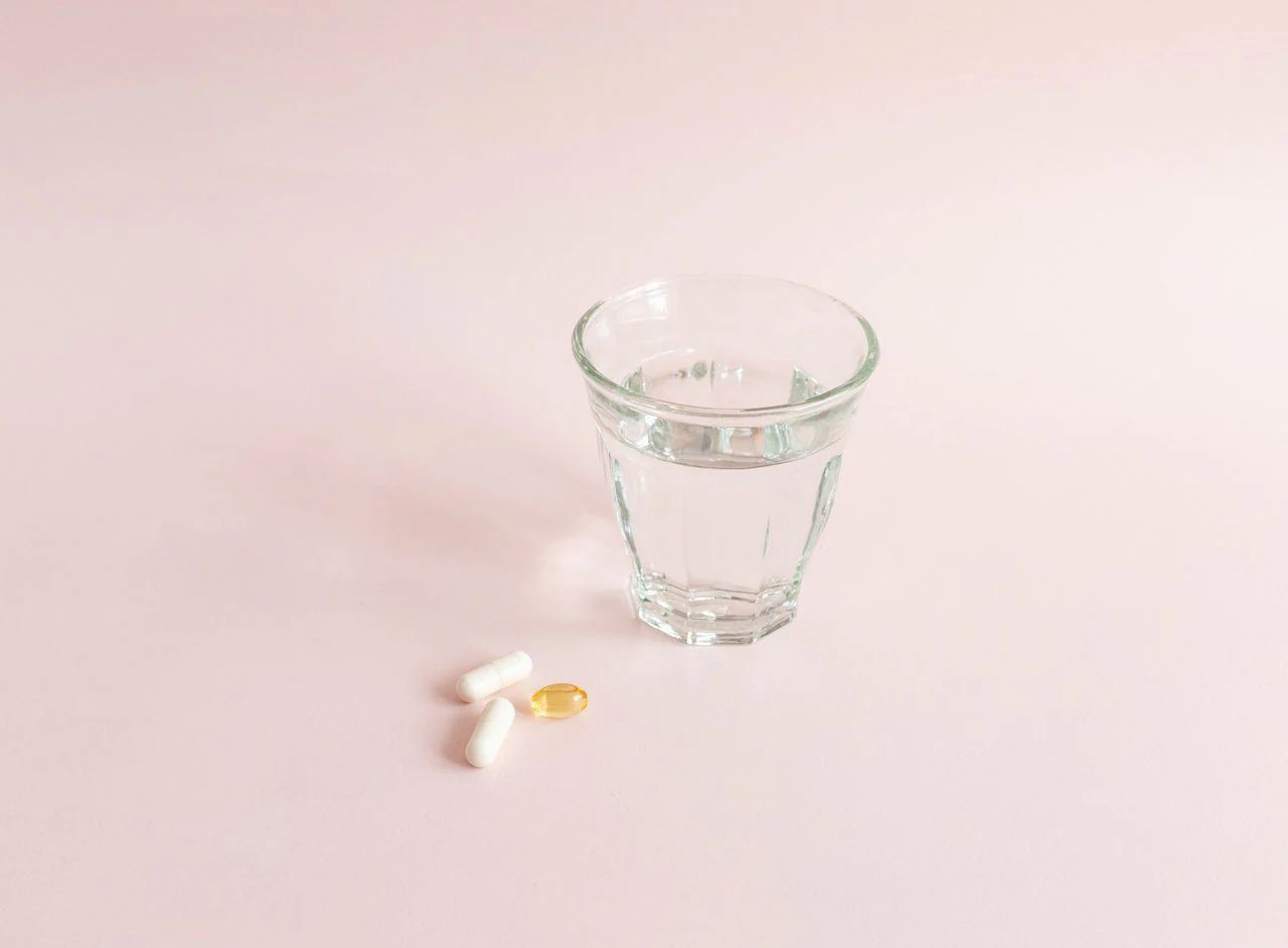 Glas Wasser, daneben zwei Kapseln und eine Kapsel. Auf rosa Untergrund.