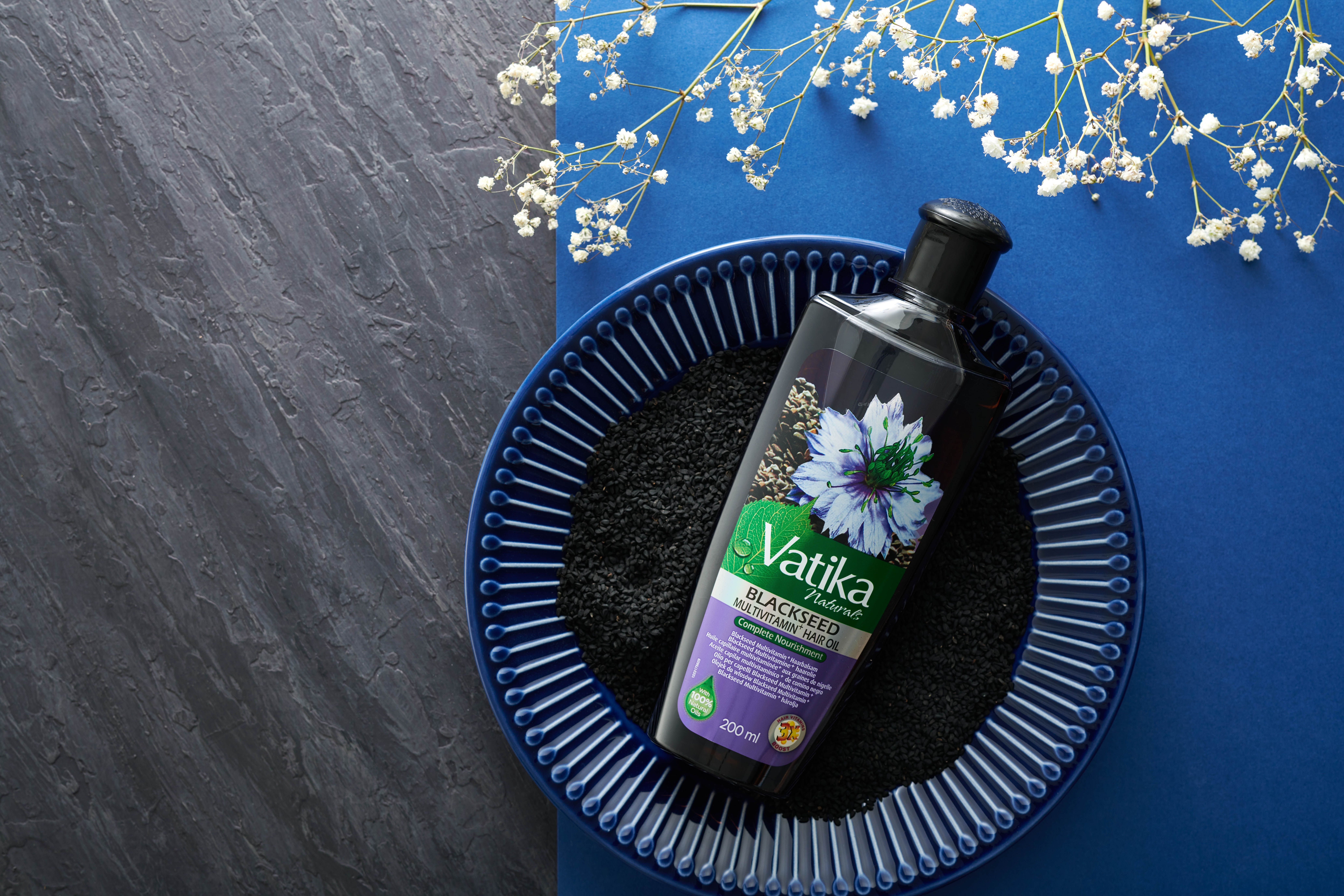 Vatika Naturals Schwarzkümmel Haaröl auf blauem Teller mit Schwarzkümmel. Auf blauem und grauem Untergrund.