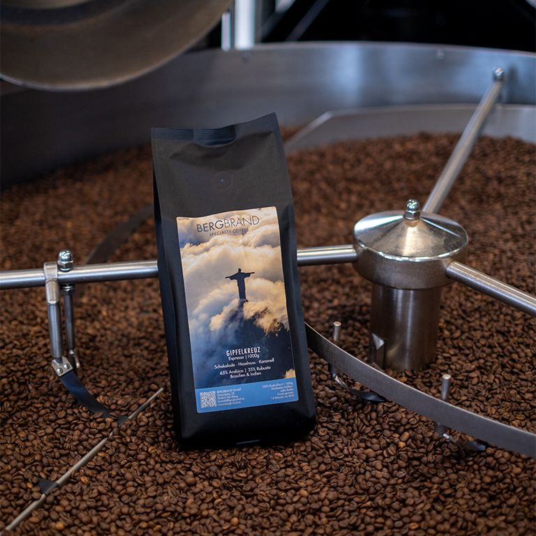 Schwarzer Beutel mit BERGBRAND Gipfelkreuz Kaffee. Aufschrift und Bild des Cristo Redentor. Kaffeebohnen im Hintergrund.