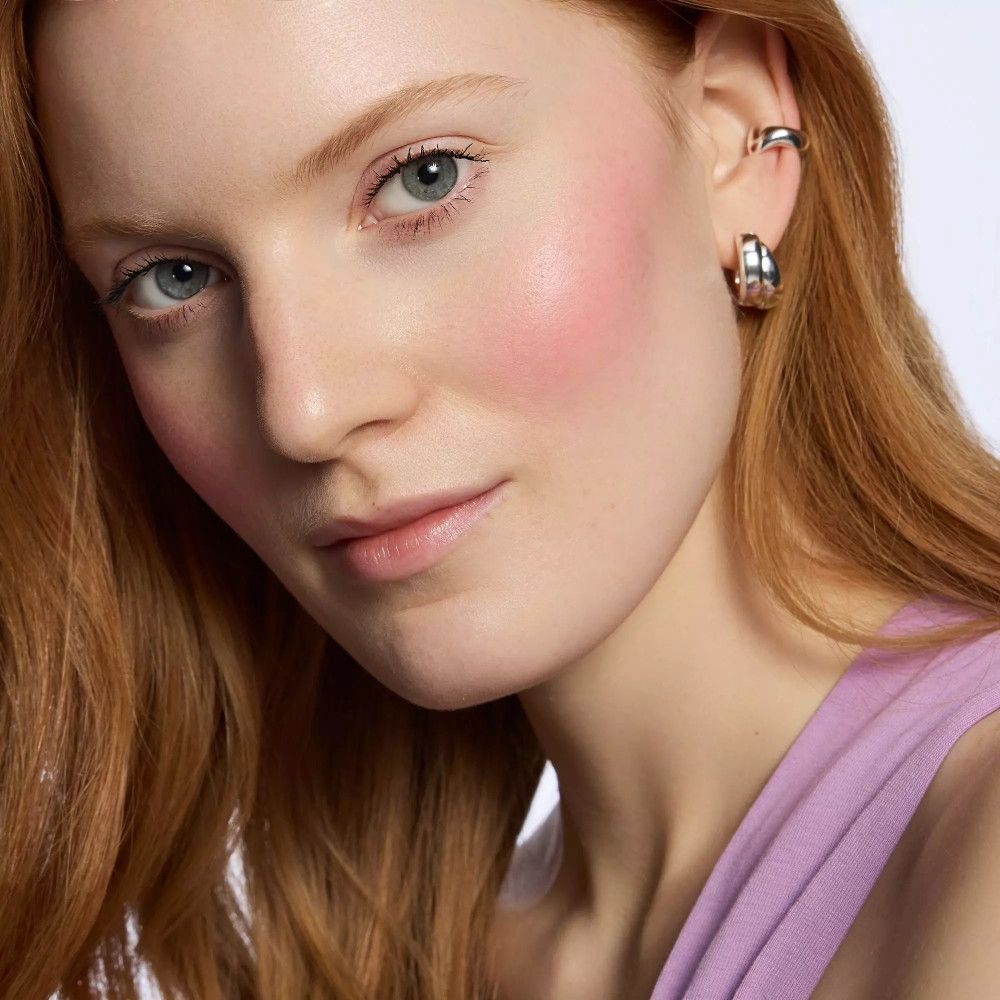 Porträt einer Frau mit Rouge auf den Wangen. Sie trägt Ohrringe. Rotes Haar. Heller Hintergrund.