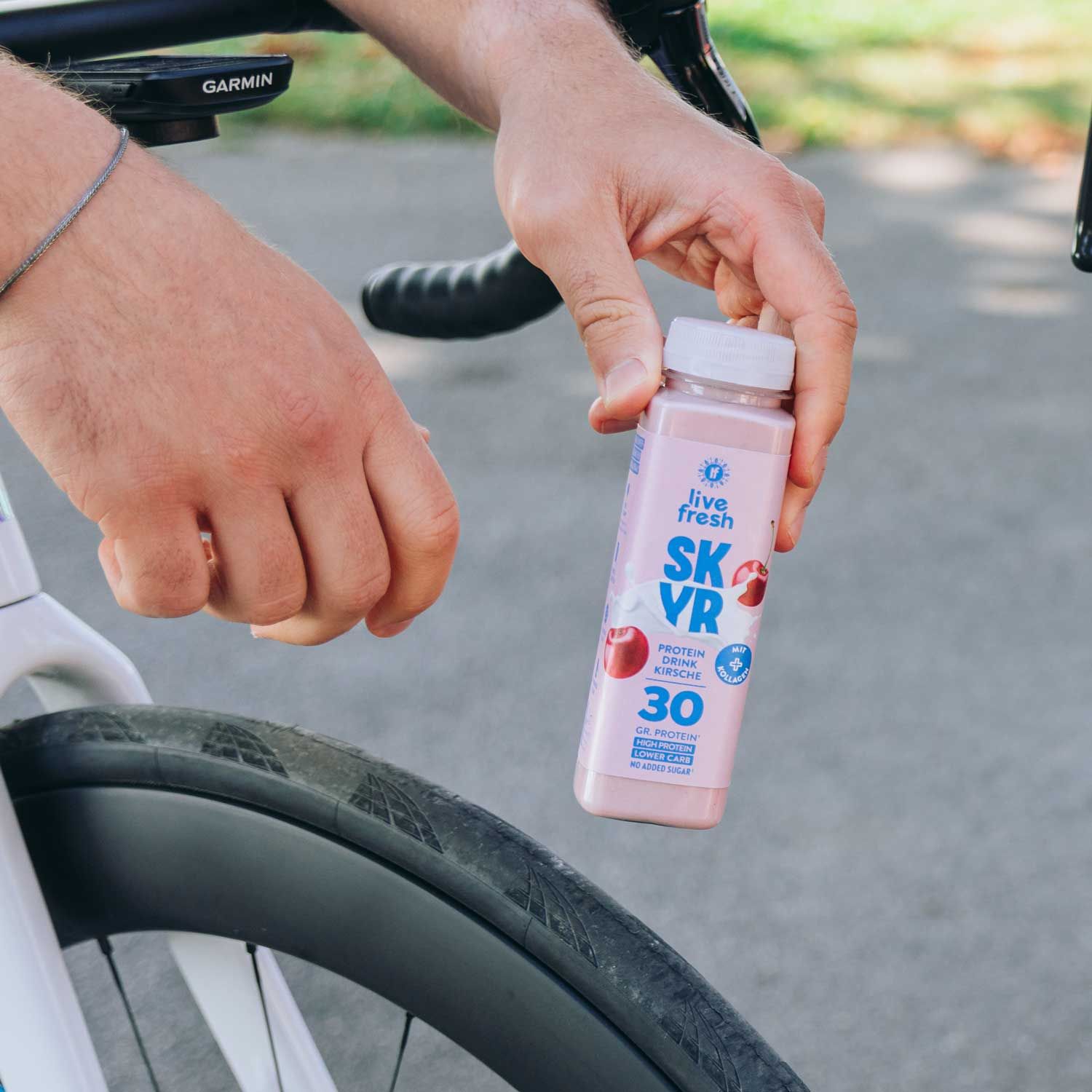 Person hält eine Flasche Skyr Drink Kirsche neben einem Fahrrad. Aufdruck: Skyr, Kirsche, 30g Protein, hoher Proteingehalt, wenig Zucker.