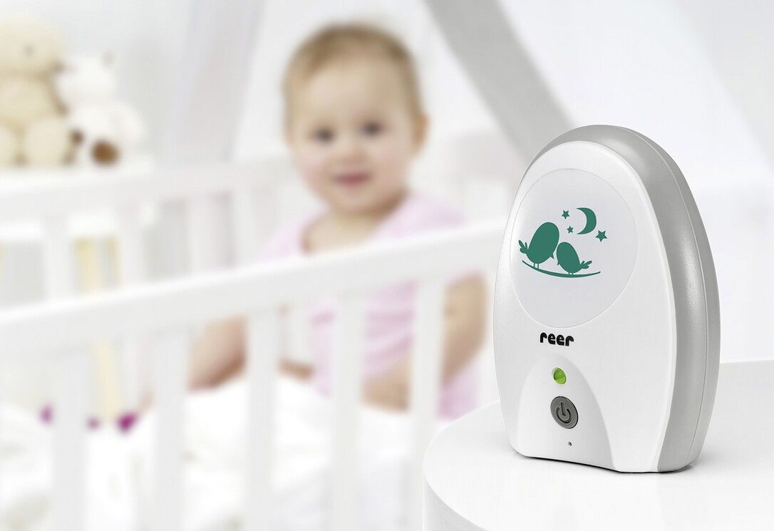 Babyphone Neo, weiß-grau. Vögel-Motiv auf der Vorderseite. Ein-/Ausschalter und grüne LED. Steht auf einem Tisch neben einem Babybett. Marke: reer.