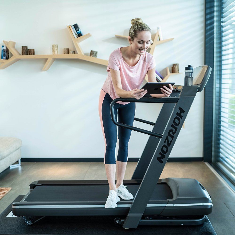 Frau steht an einem Laufband und hält ein Tablet. Laufband mit Horizon-Logo. Frau trägt Sportkleidung.
