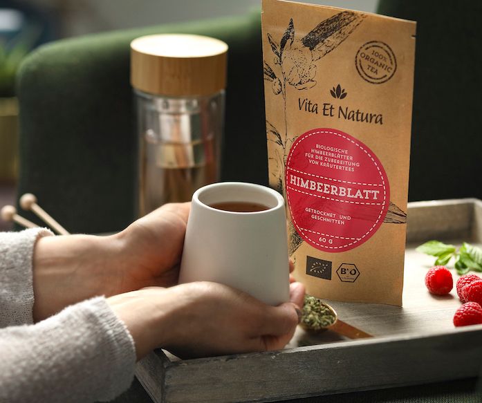 Person hält eine Tasse Tee. Daneben Packung Himbeerblätter, Glas mit Tee und frische Himbeeren. Aufdruck: Vita Et Natura, 100% Organic Tea.