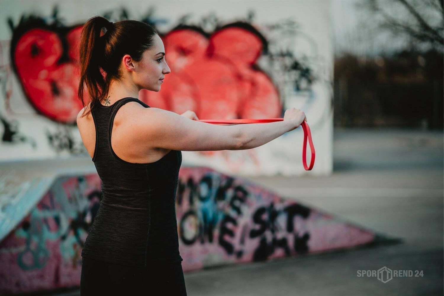 Frau trainiert mit rotem Fitnessband im Freien. Sie steht seitlich vor einer Graffiti-Wand. Das Band ist um ihre Arme gelegt.