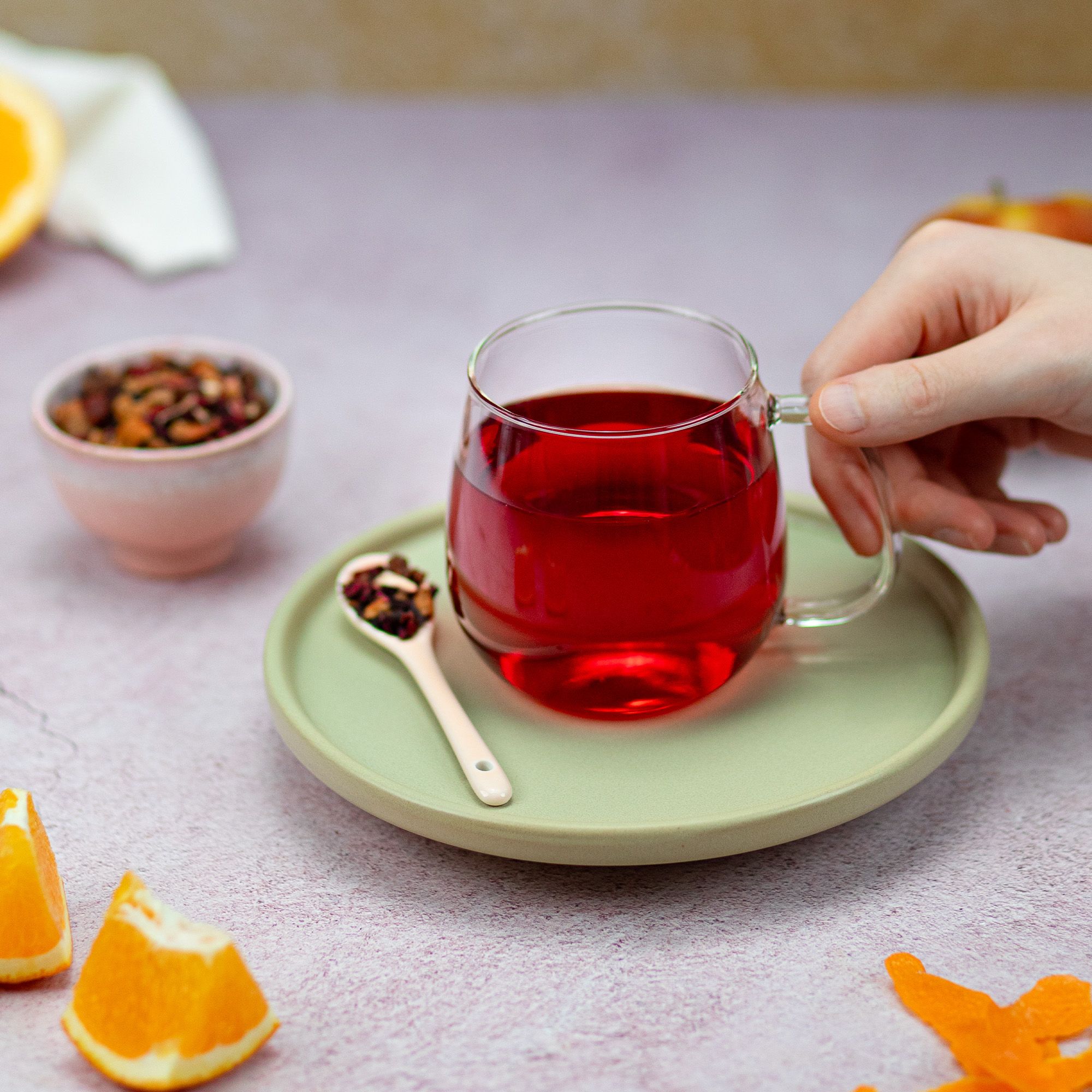 Glasbecher mit rotem Getränk auf Untertasse. Hand hält Becher. Löffel mit Tee daneben. Orangenstücke.