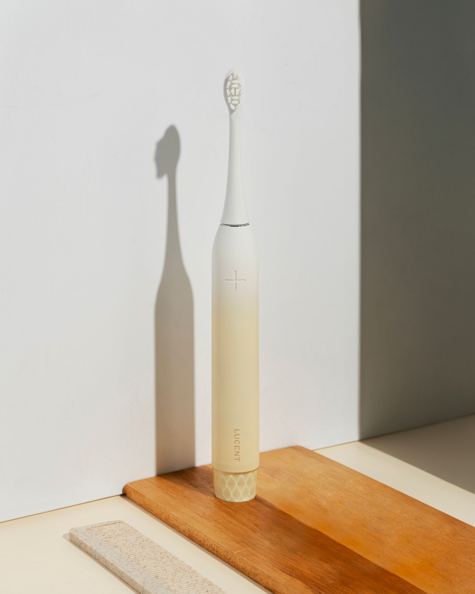 Vanillefarbene elektrische Zahnbürste. Bürstenkopf weiß. Aufdruck: LUCENT. Liegt auf Holzbrett.