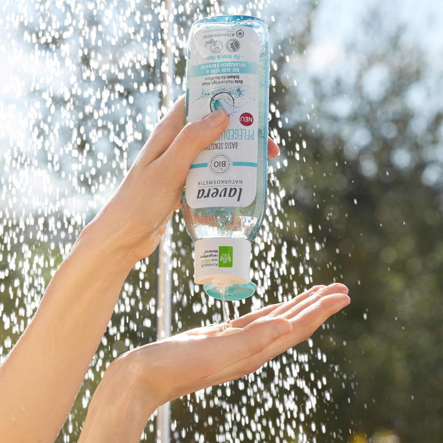 Hände halten hellblaue Flasche. Produkt: lavera Basis Sensitiv Pflegedusche 2in1. Wasserstrahlen im Hintergrund.