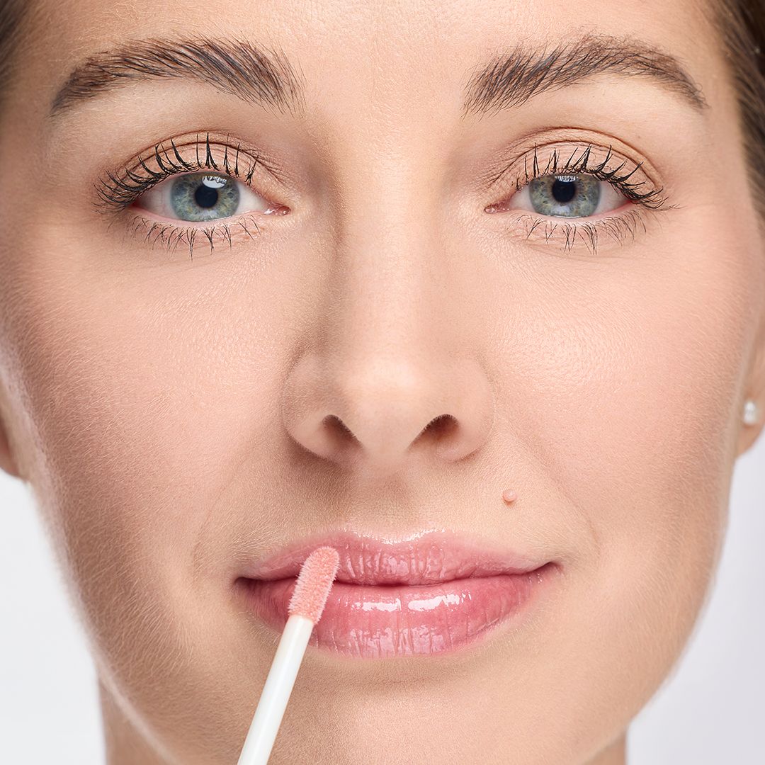 Nahaufnahme von Lippen mit Lipgloss. Ein Applikator wird verwendet. Der Lipgloss ist rosa und glänzend.