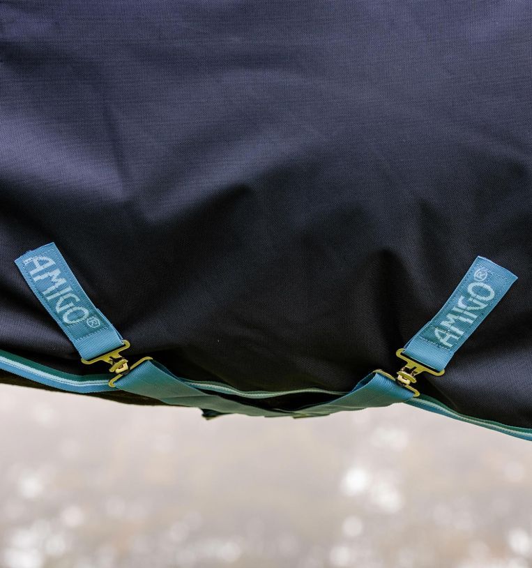 Nahaufnahme der Unterseite einer dunkelblauen Pferdedecke. Zwei hellblaue Riemen mit goldenen Schnallen. 'AMIGO' Schriftzug auf den Riemen.