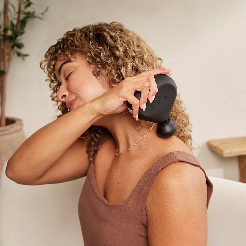 Frau verwendet ein schwarzes Therabody Vibrationsmassagegerät am Nacken. Braunes Oberteil. Goldene Halskette. Helle Umgebung.