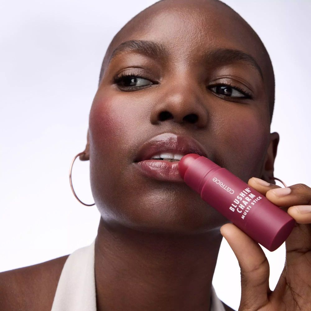Frau mit Rouge auf den Wangen und Lippen. Hält einen Catrice Blush Stick. Dunkle Haut.