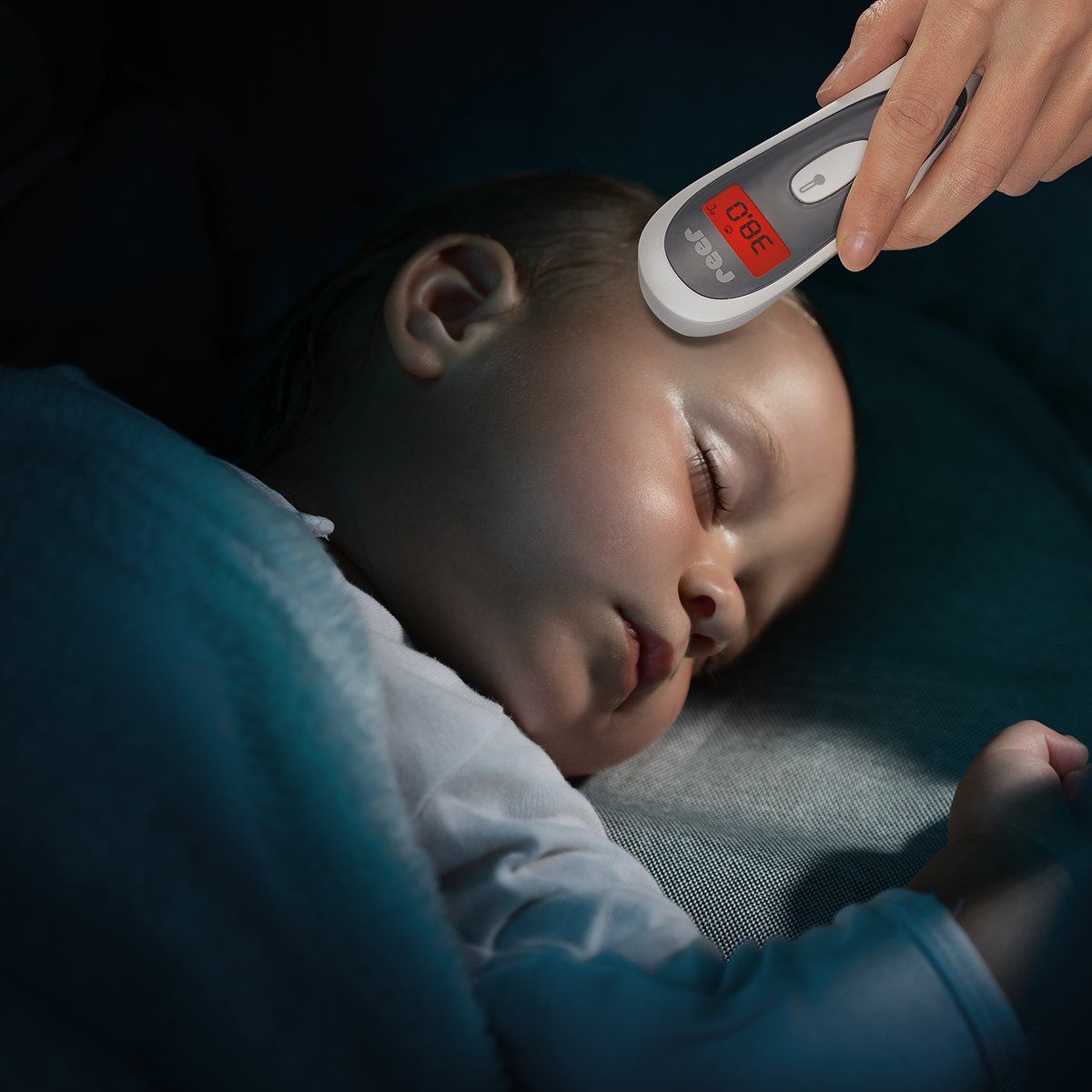 Hand hält ein kontaktloses Infrarot-Thermometer an die Stirn eines schlafenden Babys. Anzeige: 38,0 °C.