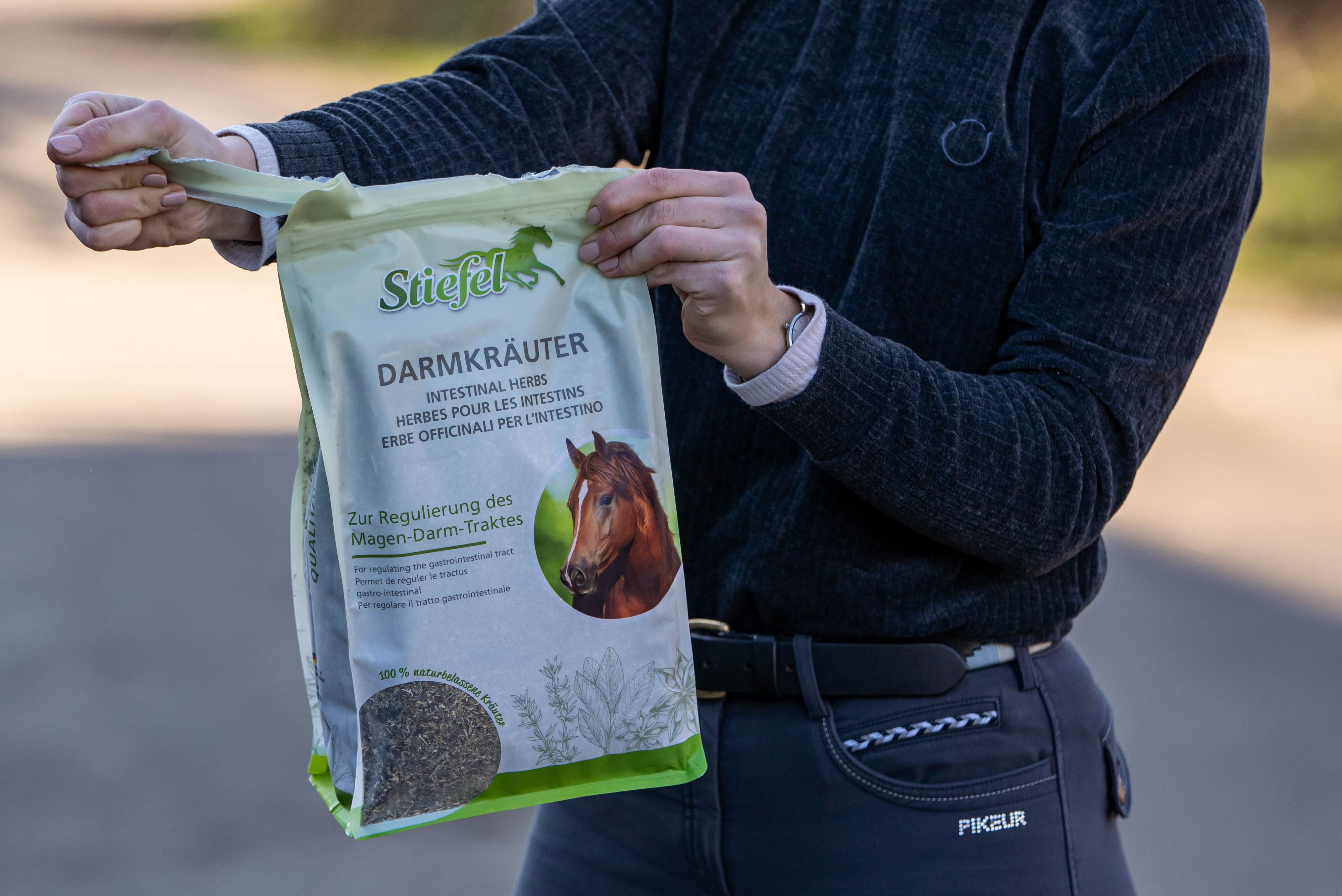 Person hält einen Beutel Stiefel Darmkräuter. Der Beutel ist grün-weiß und zeigt ein Pferd. Aufschrift: Darmkräuter, Intestinal Herbs, Erbe Officinali per l'intestino.