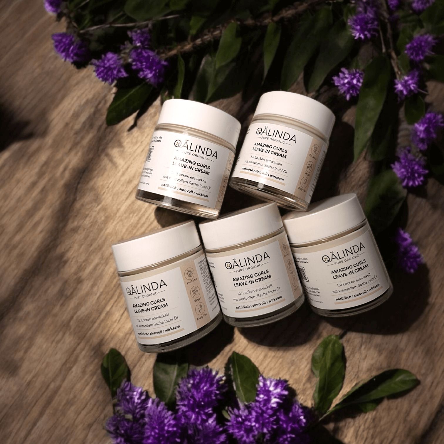Fünf Tiegel QAELINDA Amazing Curls Leave-In Cream. Beige Tiegel mit weißem Deckel. Text auf Etikett. Auf Holz mit lila Blüten und Blättern.