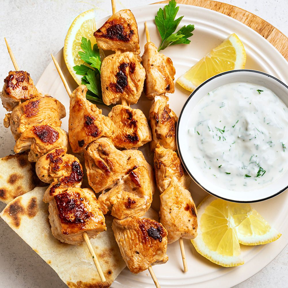 Teller mit Hähnchenspießen, Tzatziki, Zitronen und Fladenbrot. Holzbrett. Draufsicht.