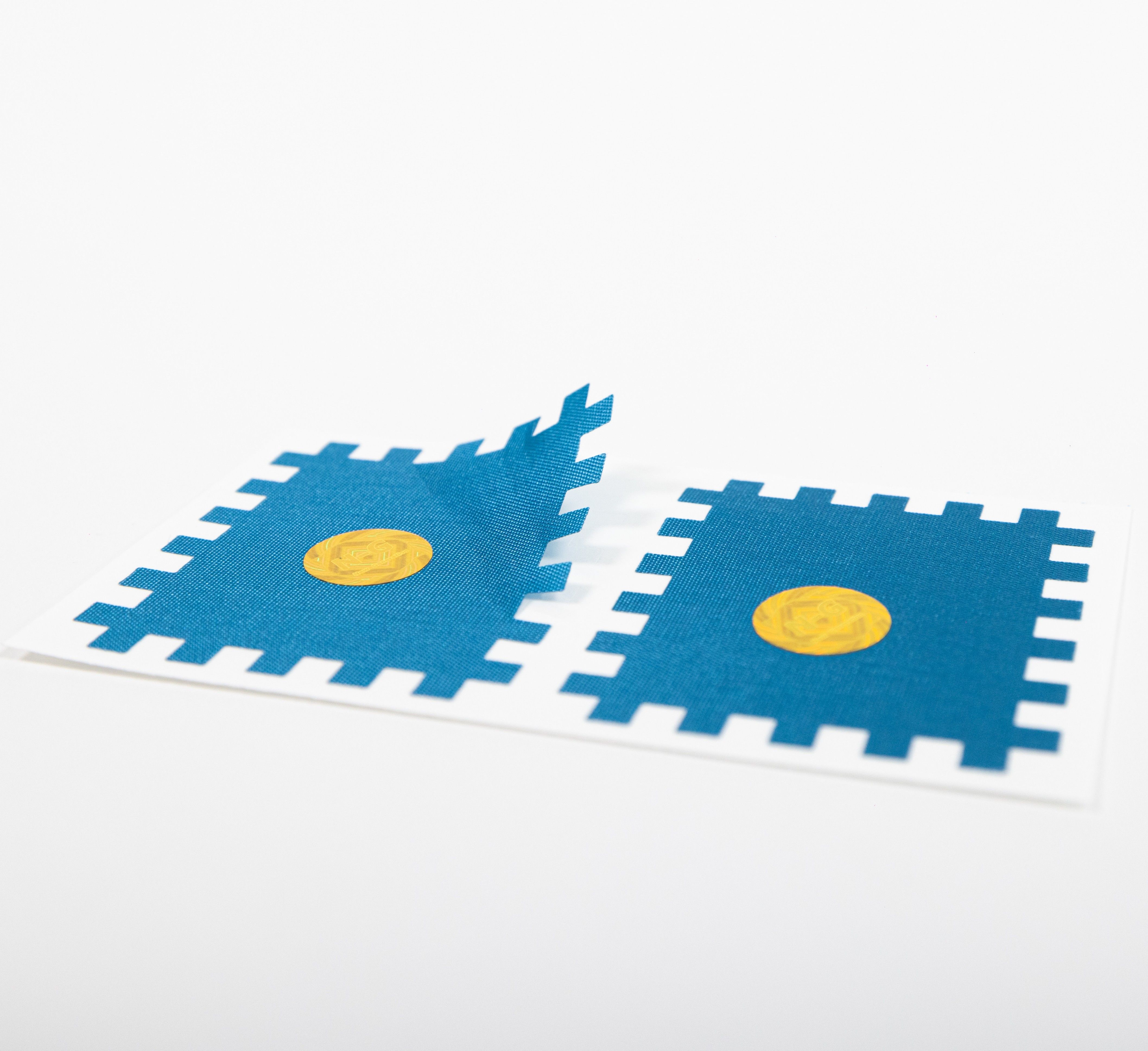 Zwei blaue, quadratische Pflaster mit gelbem Kreis in der Mitte. Pflaster mit gezackten Rändern. Auf weißem Hintergrund.