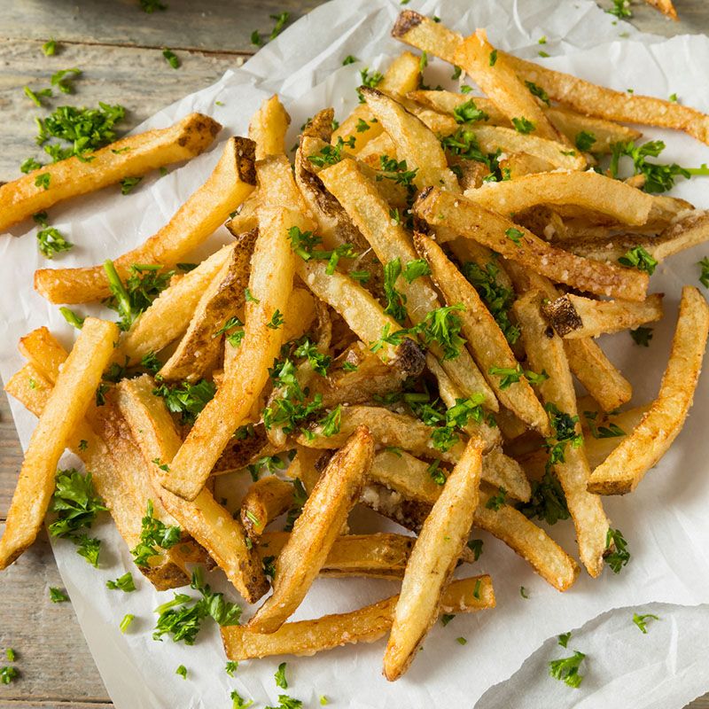 Pommes Frites auf Papier, mit Kräutern bestreut. Goldbraune Farbe, leicht gebräunt. Auf hellem Holzuntergrund.