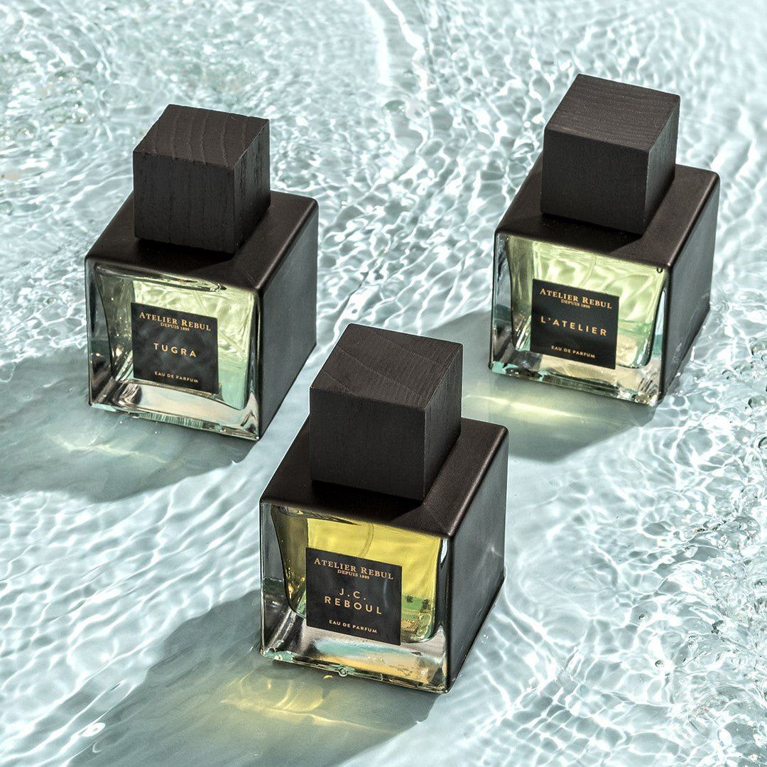 Drei Parfümflakons mit schwarzen Deckeln. Aufschrift: Tugra, L'Atelier, J.C. Rebul. Atelier Rebul Logo. Auf Wasser.