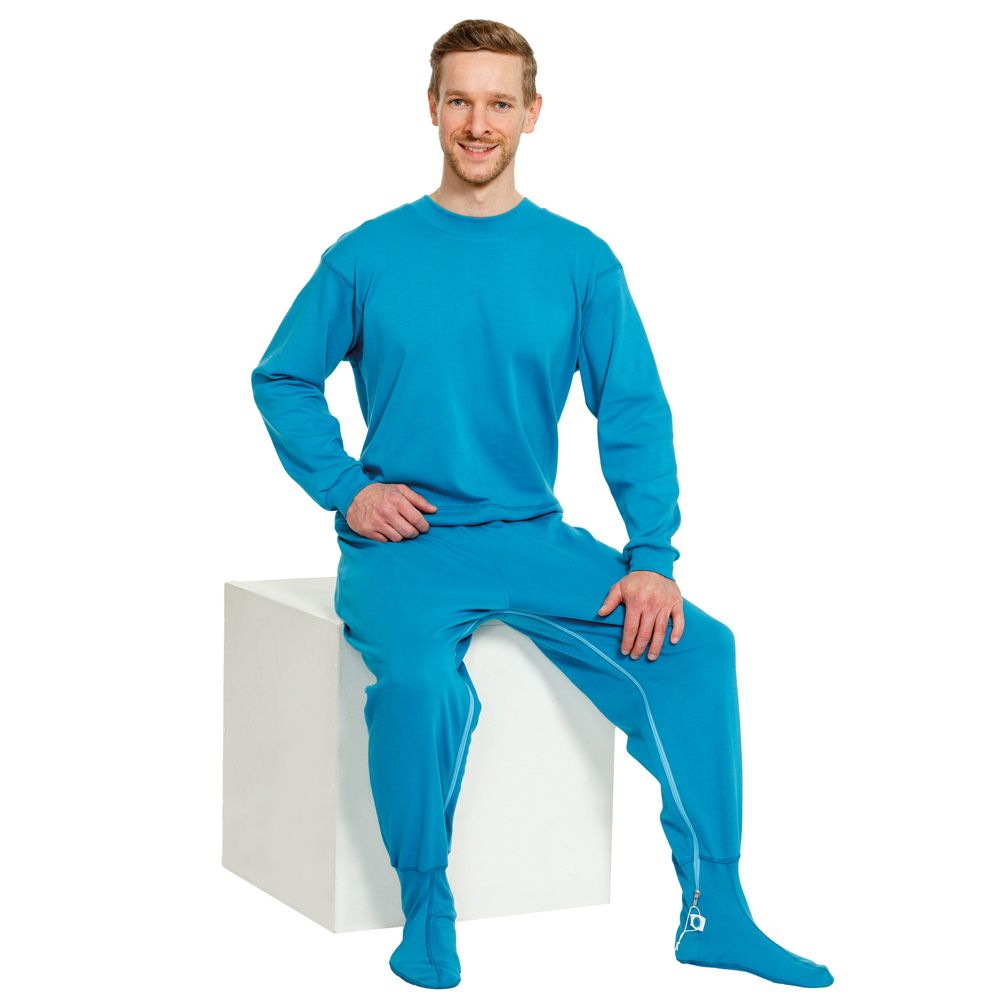 Blauer Pflegeoverall mit Fußteil. Mann sitzt auf weißem Würfel. Reißverschluss an den Beinen.