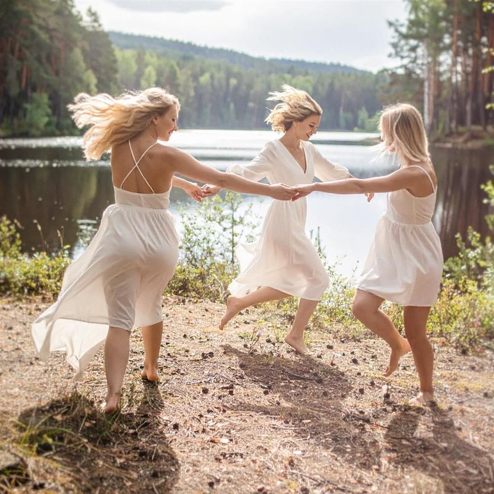 Drei Frauen tanzen im Freien am Seeufer. Sie tragen weiße Kleider und halten sich an den Händen. Der Hintergrund zeigt einen See und Bäume.