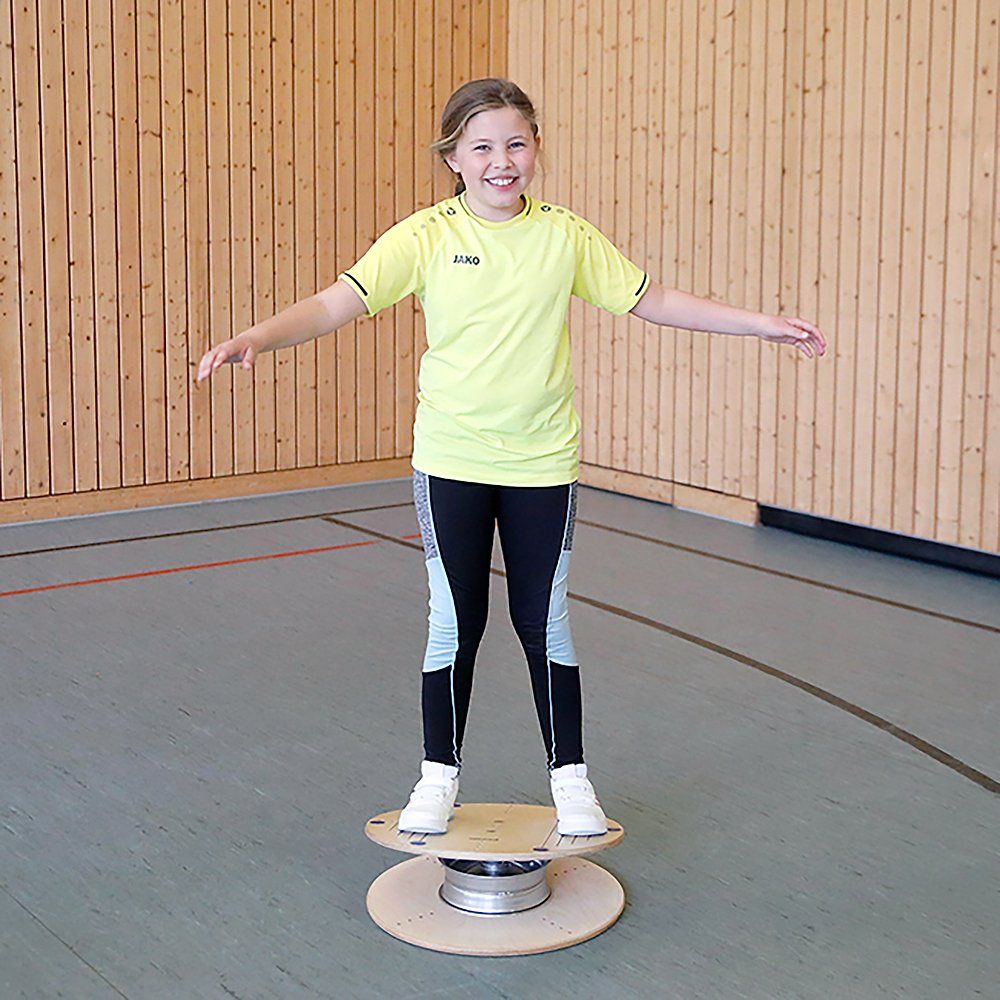 Kind steht auf einem Pedalo Gleichgewichtstrainer in einer Sporthalle. Das Gerät besteht aus Holz und Metall.