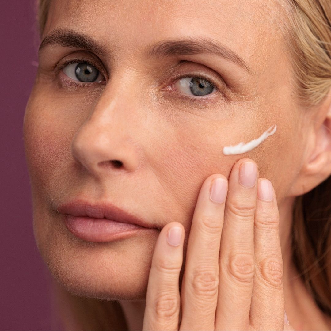 Nahaufnahme eines Gesichts. Eine Hand trägt Creme auf die Haut auf. Sichtbare Creme auf der Haut. Hintergrund: violett.