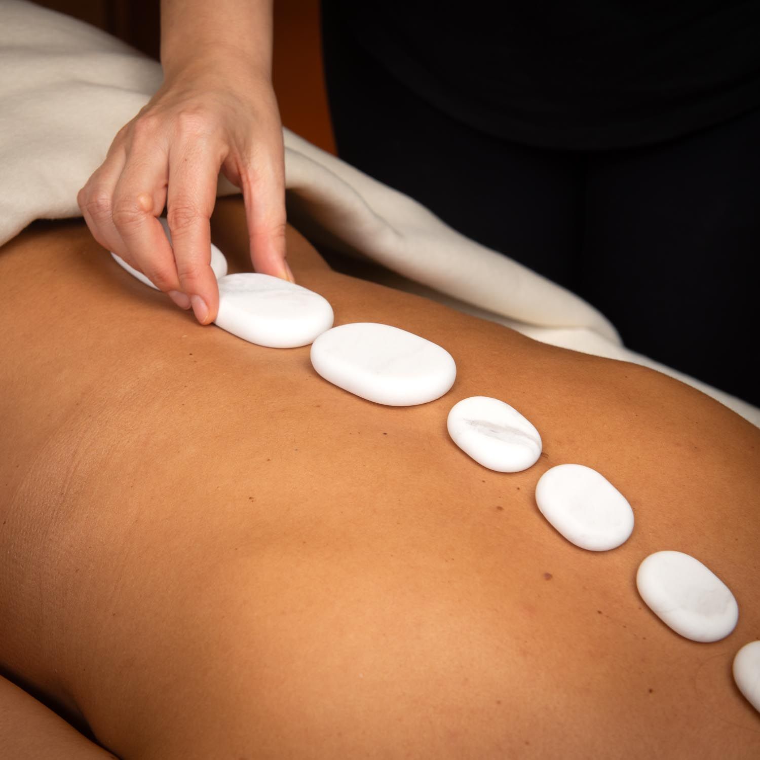 Person legt weiße, ovale Steine auf den Rücken einer Person. Auf einer Liege.