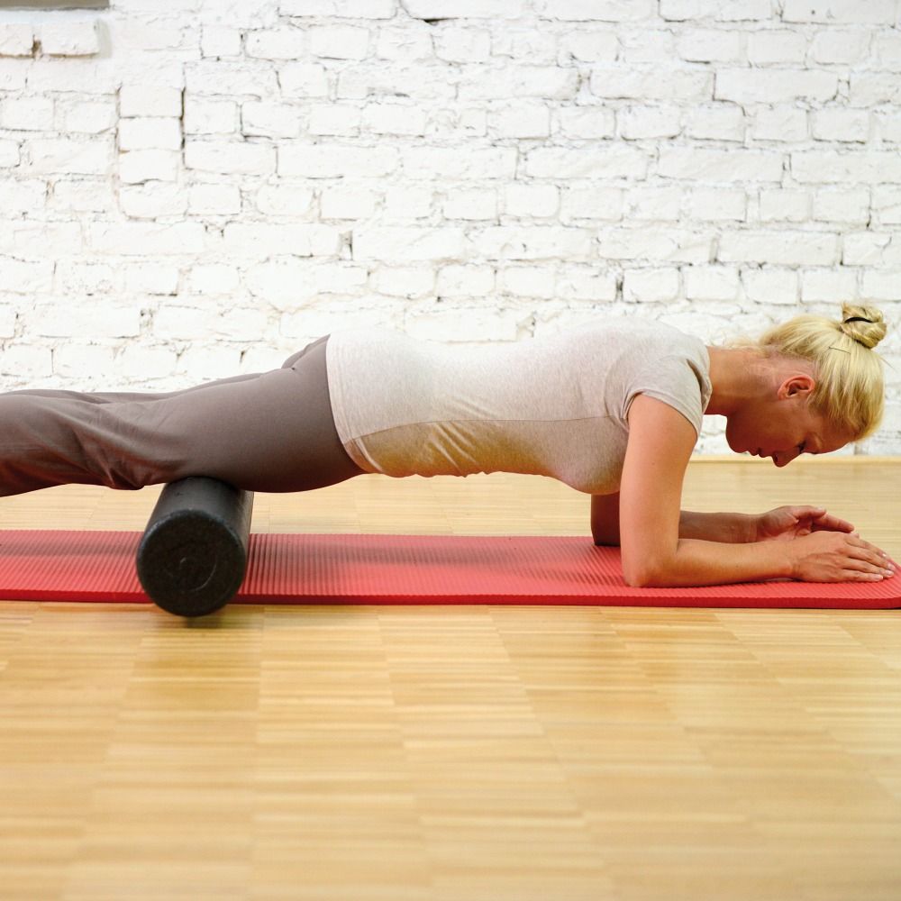 Frau auf roter Matte, stützt sich auf Unterarme. Schwarze Rolle unter Oberschenkeln. Hintergrund: Backsteinmauer.