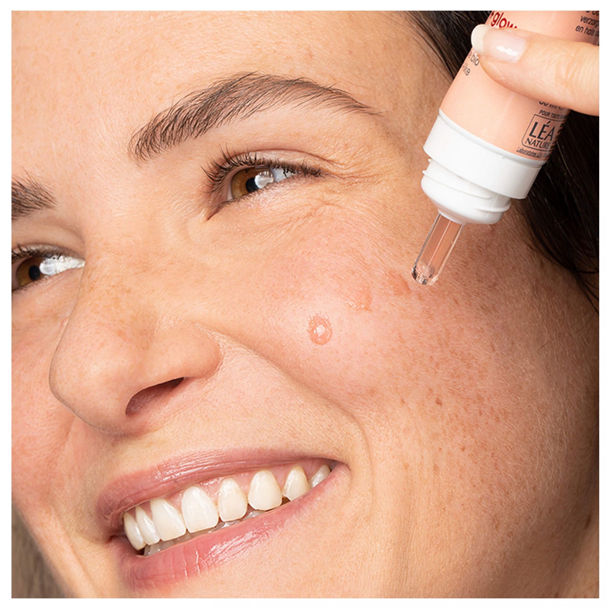 Eine Hand trägt einen Tropfer mit Serum auf die Wange einer lächelnden Frau auf. Ein Tropfen Serum ist sichtbar.