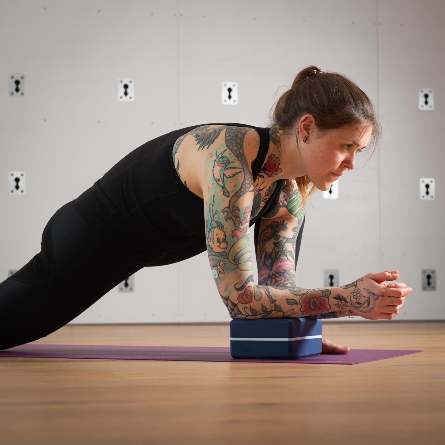 Frau in Plank-Position, Arme auf blauem Yogablock. Auf lilafarbener Matte. Tattoo-Arme.