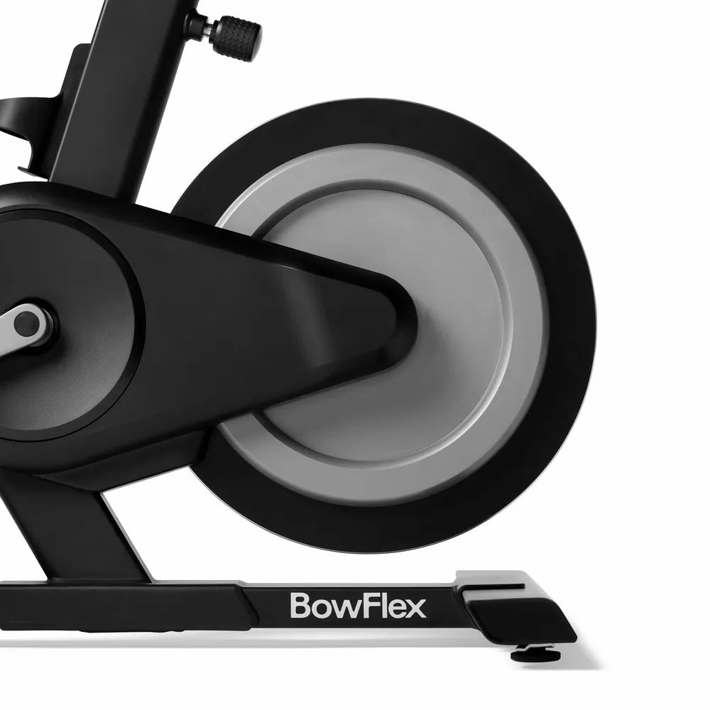 Gros plan du vélo d'intérieur. Cadre noir, volant d'inertie gris. Logo BowFlex. Pédales à sangles.