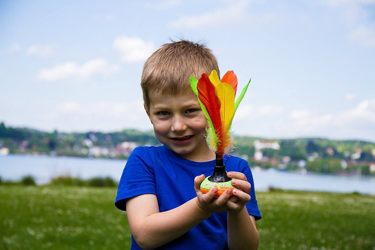 Junge hält Peteca-Shuttle. Gelbe, grüne und orangefarbene Federn. Kind trägt blaues T-Shirt.