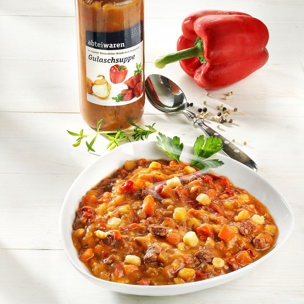 Gulaschsuppe in Schüssel, daneben Glas, Paprika, Löffel und Gewürze.