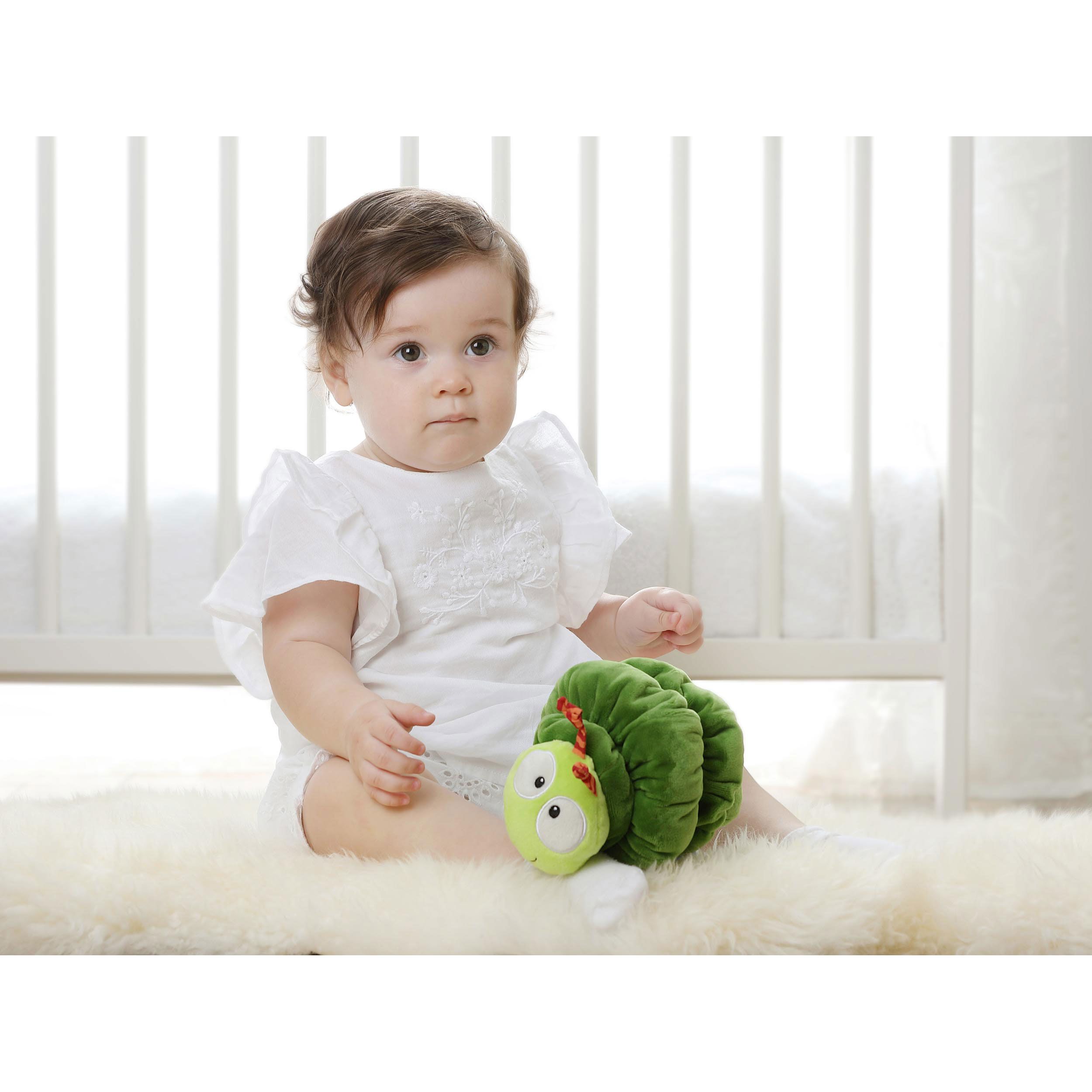 Baby sitzt mit grüner Raupe auf Fell. Raupe: gelber Kopf, Augen, Fühler.