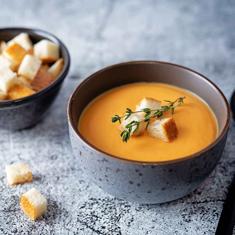 Graue Schüssel mit orangefarbener Suppe, garniert mit Croutons und Kräutern. Auf grauem Untergrund, daneben eine weitere Schüssel.