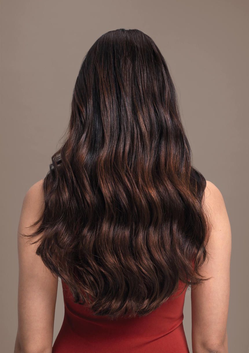 Rückansicht einer Frau mit welligem, braun gefärbtem Haar. Die Haare sind schulterlang und haben Highlights.