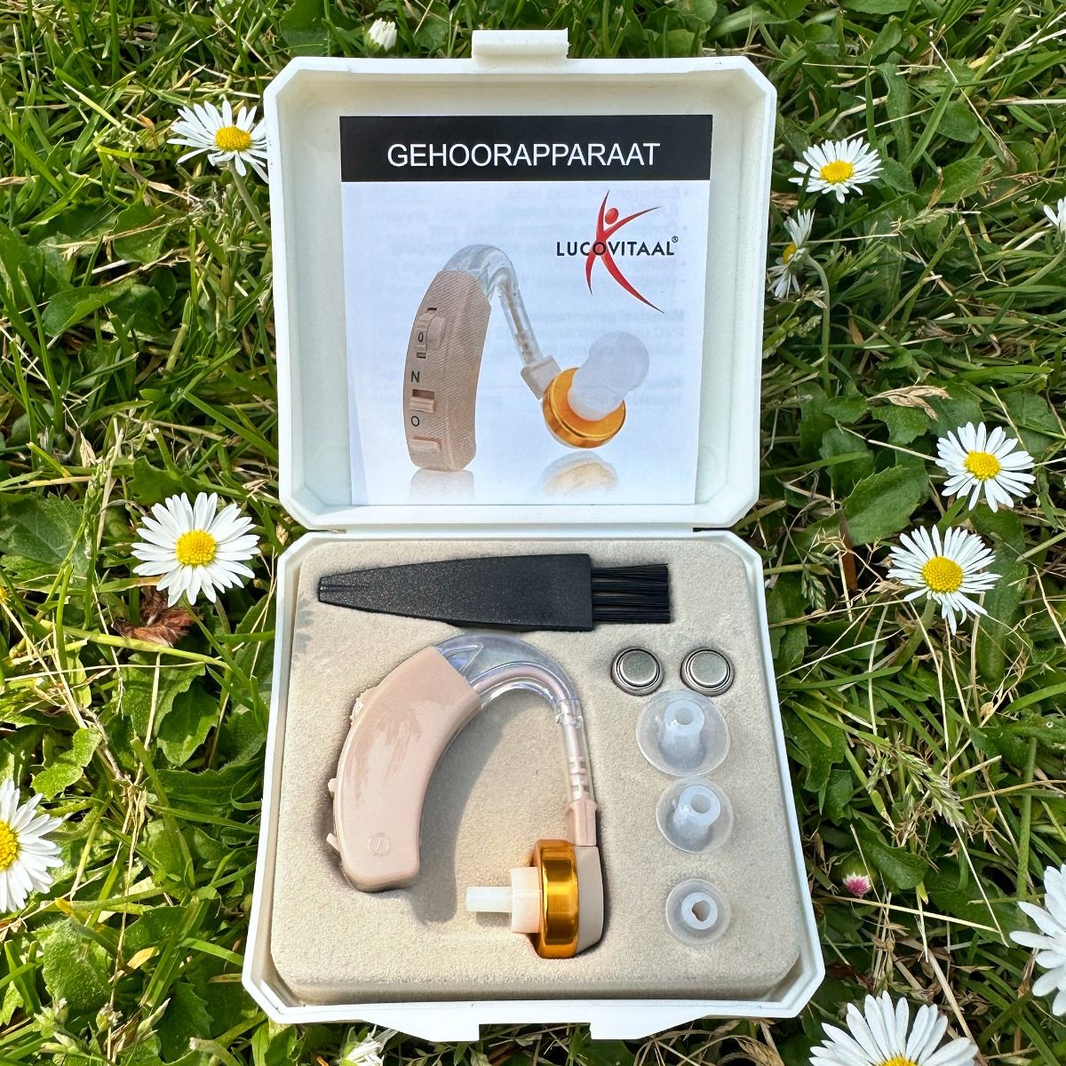 Hoortoestel in witte doos, open op gras. Bevat toestel, oordopjes, batterijen en schoonmaakborstel.