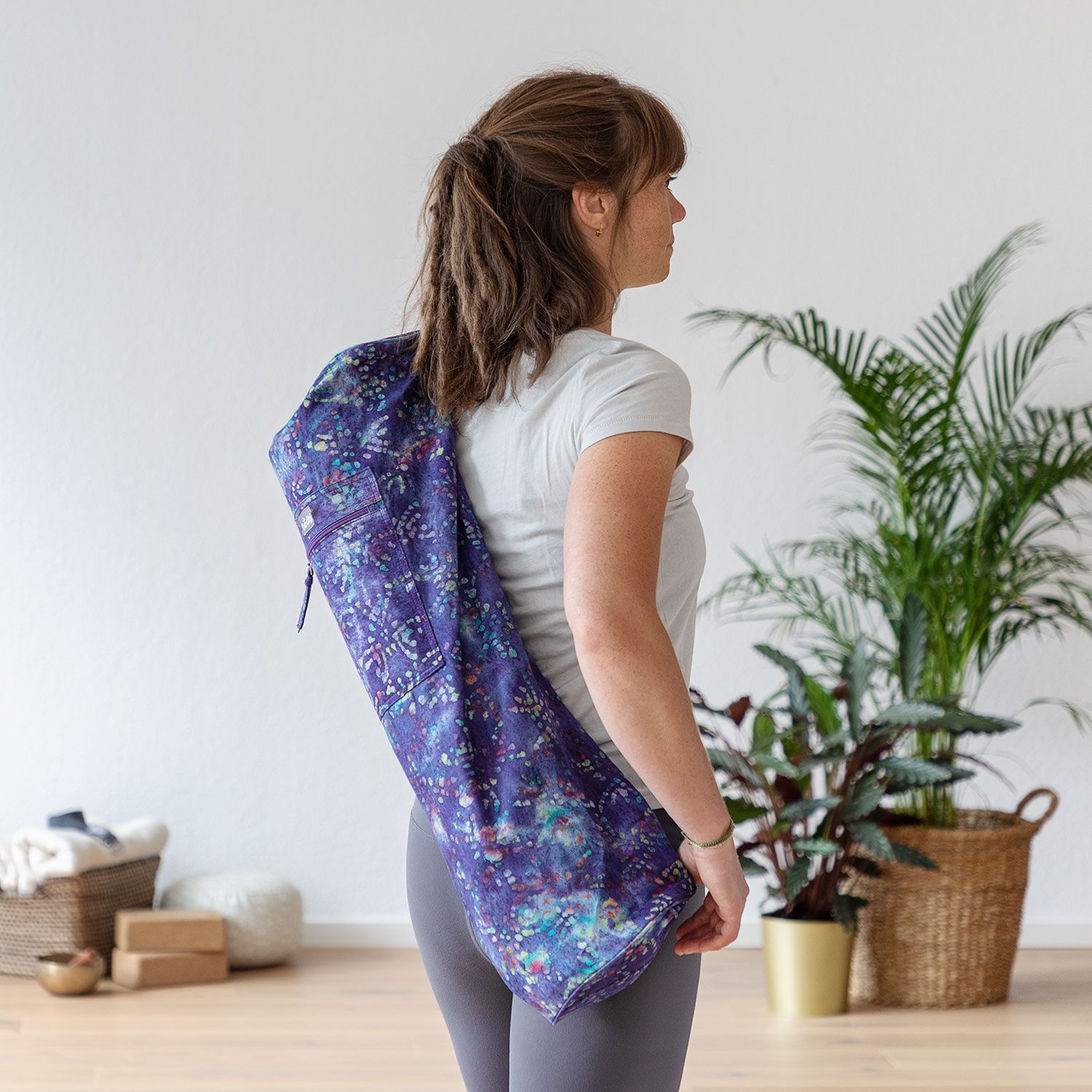 Femme portant un sac de yoga sur l'épaule. Sac lila à motifs colorés. Dans une pièce claire, avec plantes et accessoires de yoga.