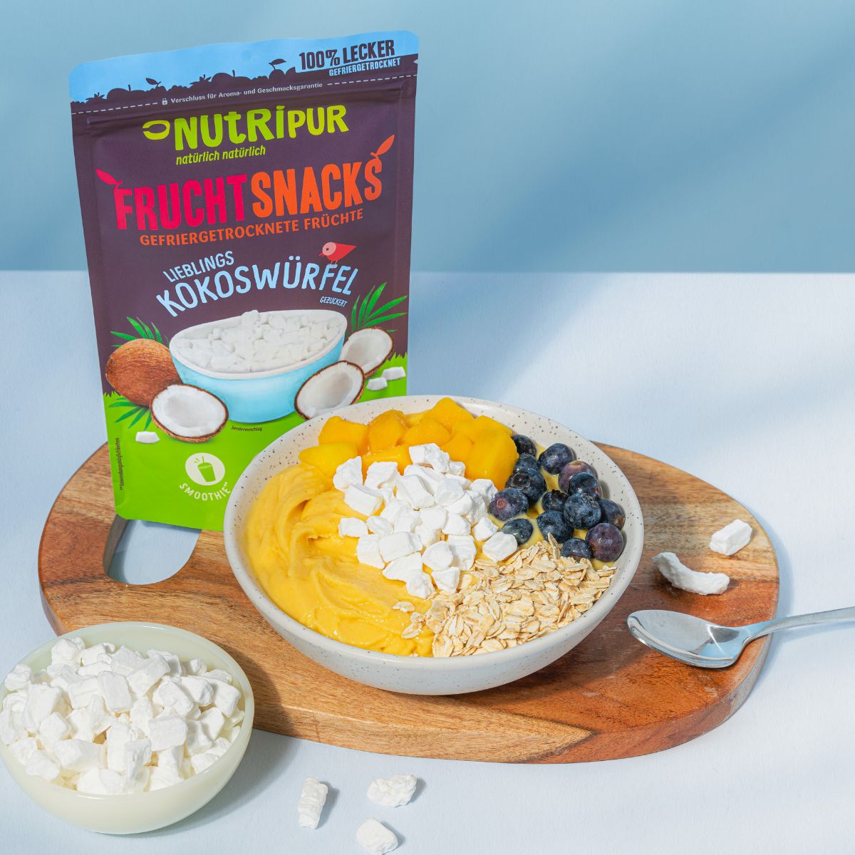 Verpackung mit NutriPur Frucht Snacks Kokoswürfel. Schüssel mit Obst, Haferflocken und Kokoswürfeln. Auf Holzbrett.