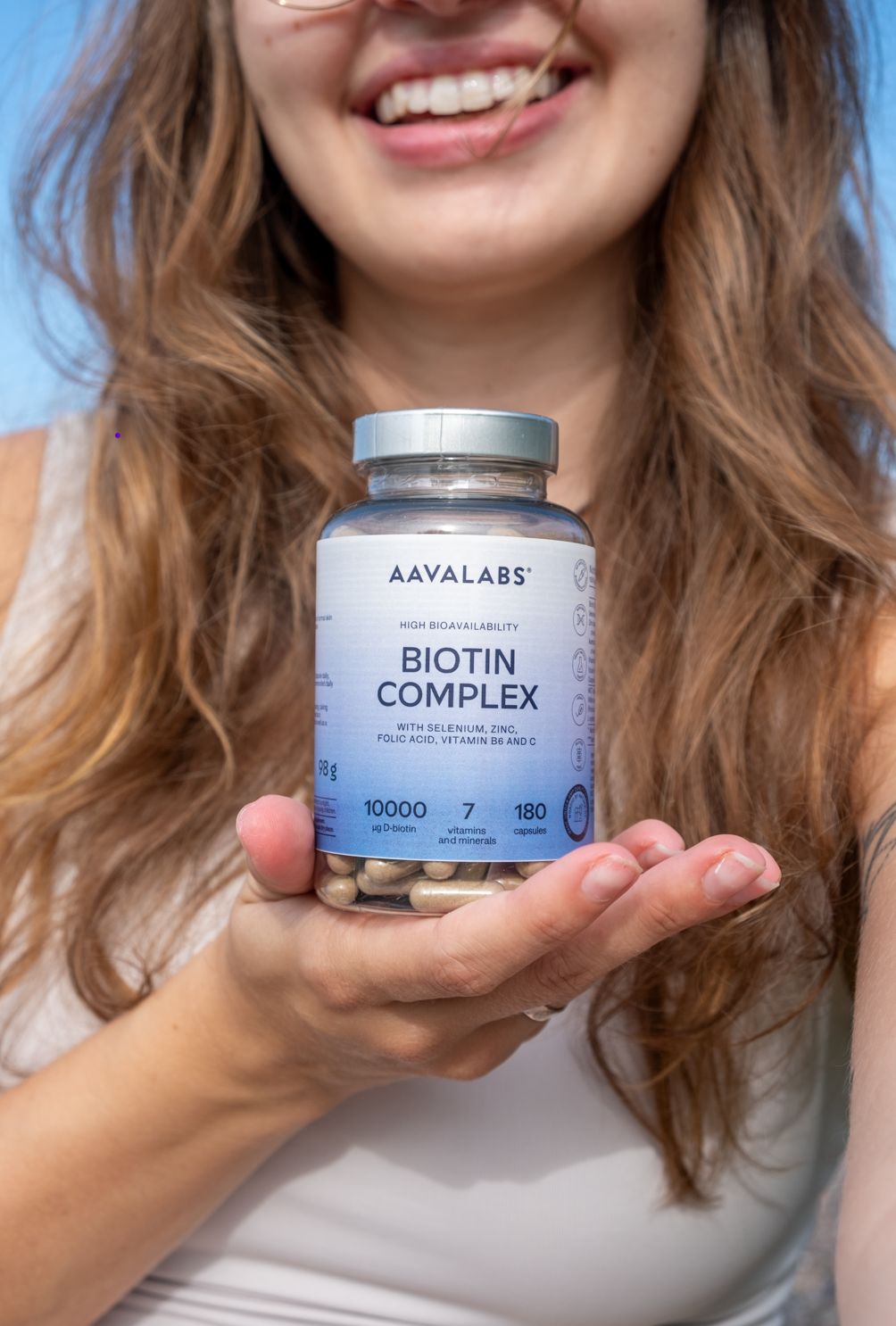 Frau hält eine Glasflasche mit Kapseln. Aufschrift: AAVALABS, Biotin Complex, 180 Kapseln. Enthält Biotin, Zink, Selen.