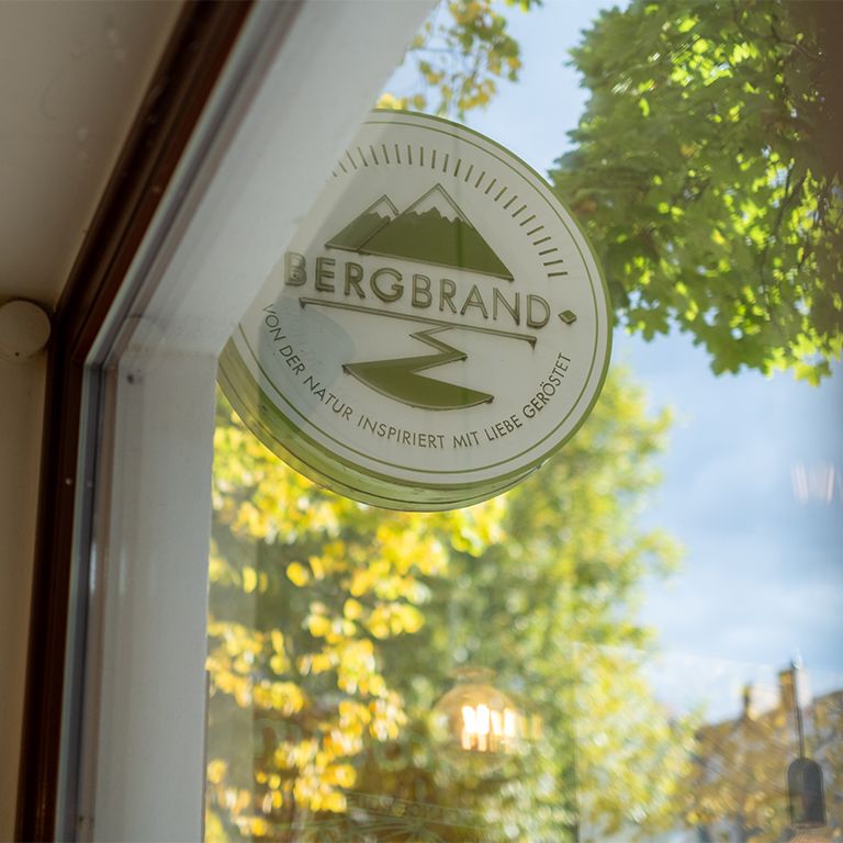 Rundes Schild mit BERGBRAND-Logo. Text: Von der Natur inspiriert mit Liebe geröstet.