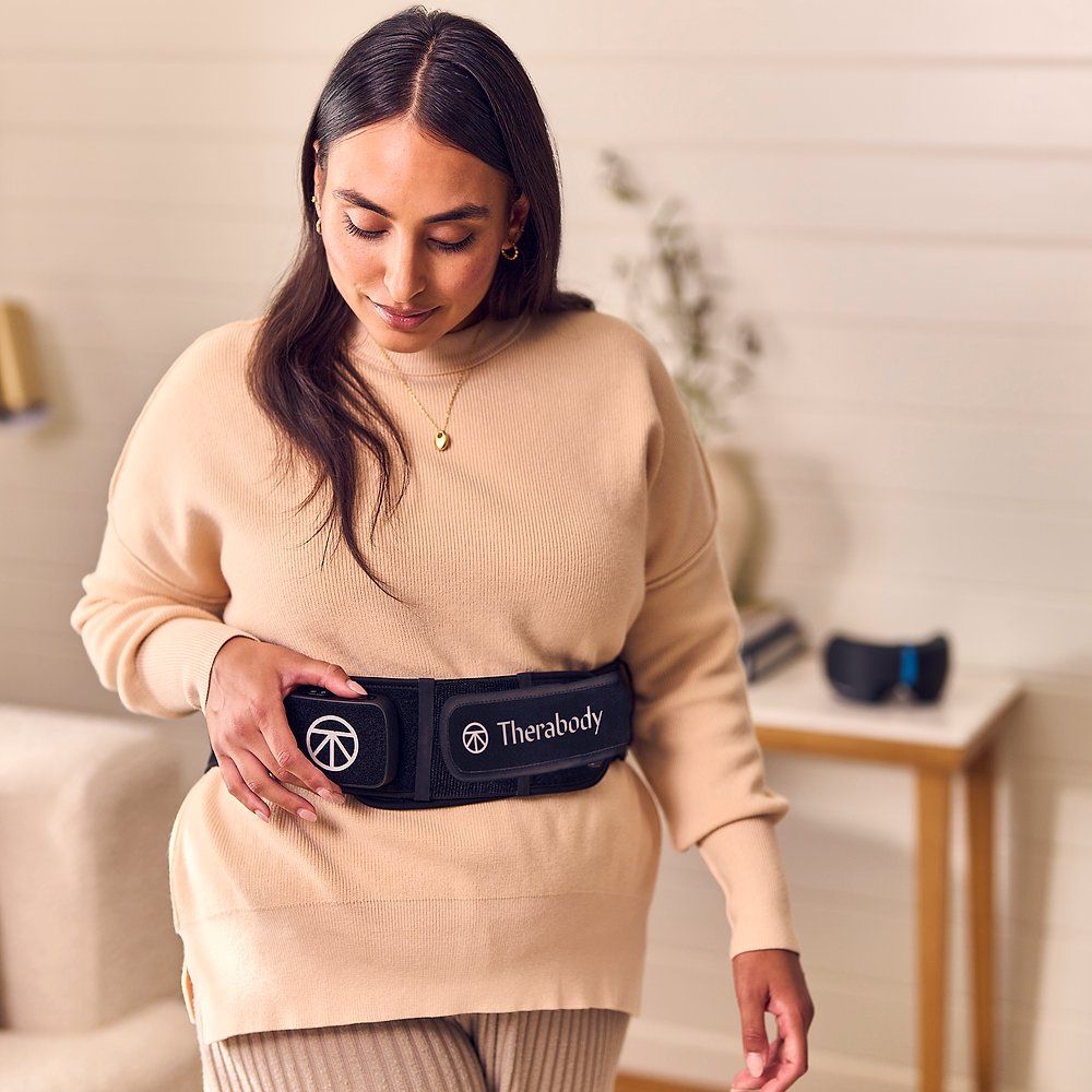 Frau trägt einen schwarzen Therabody Massagegurt. Therabody-Logo und Bedienfeld sichtbar. Beige Kleidung.