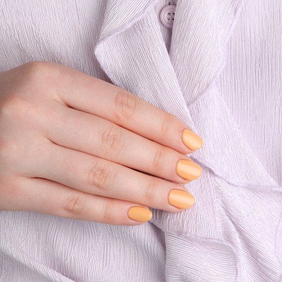 Hand mit lackierten Nägeln in Pastell-Pfirsich-Farbe. Nägel sind kurz und lackiert. Hintergrund: helles, strukturiertes Material.