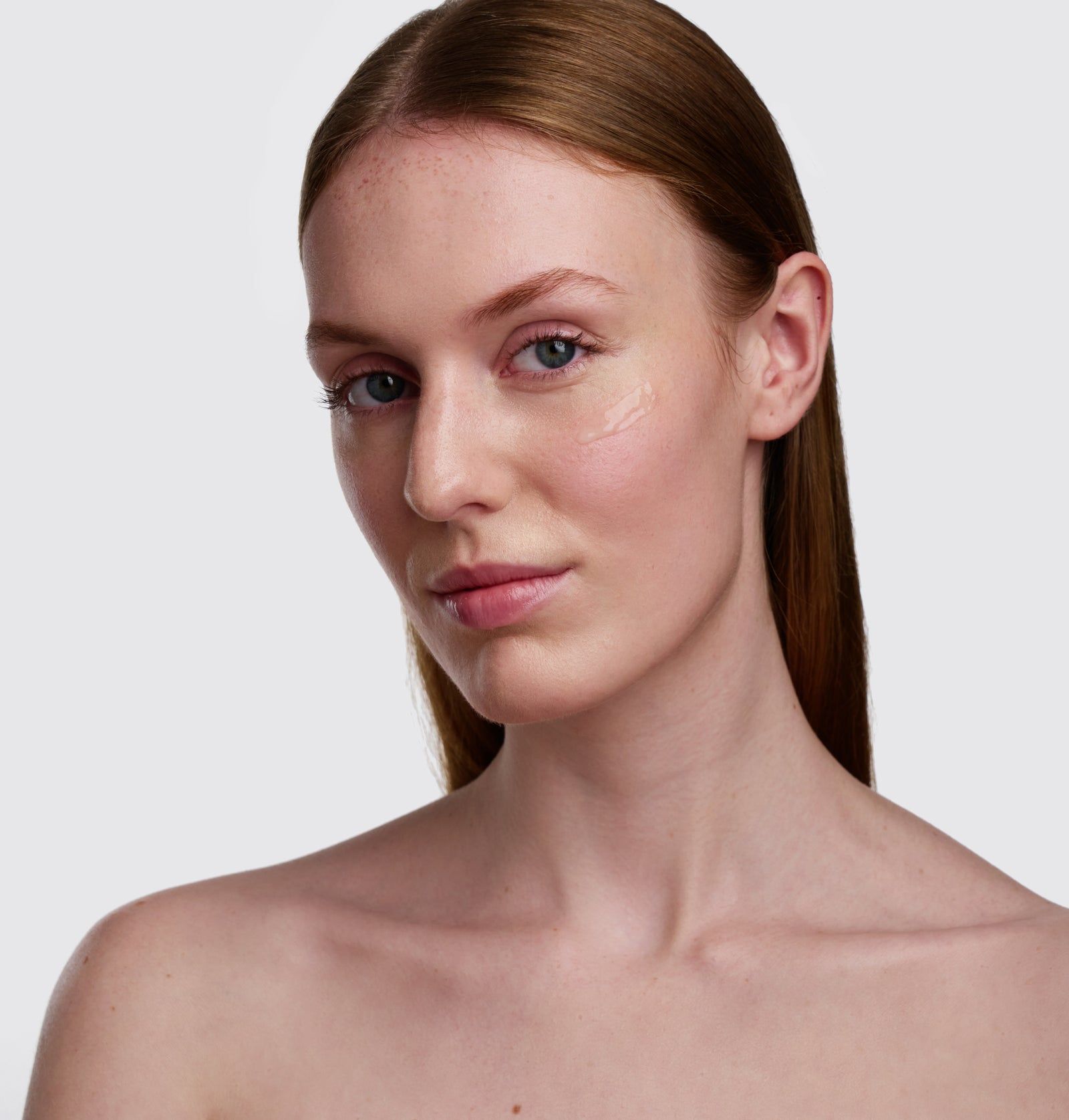 Porträt einer Frau mit Hautpflegeprodukt auf der Wange. Helle Haut, braunes Haar, Blick zur Kamera.