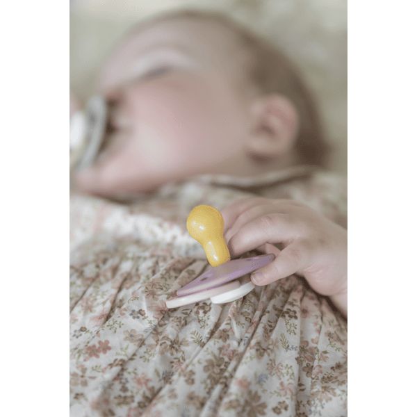 Babyhand hält Schnuller. Gelber Sauger, lila Schild. Baby liegt auf floralem Stoff. Schnuller im Mund.