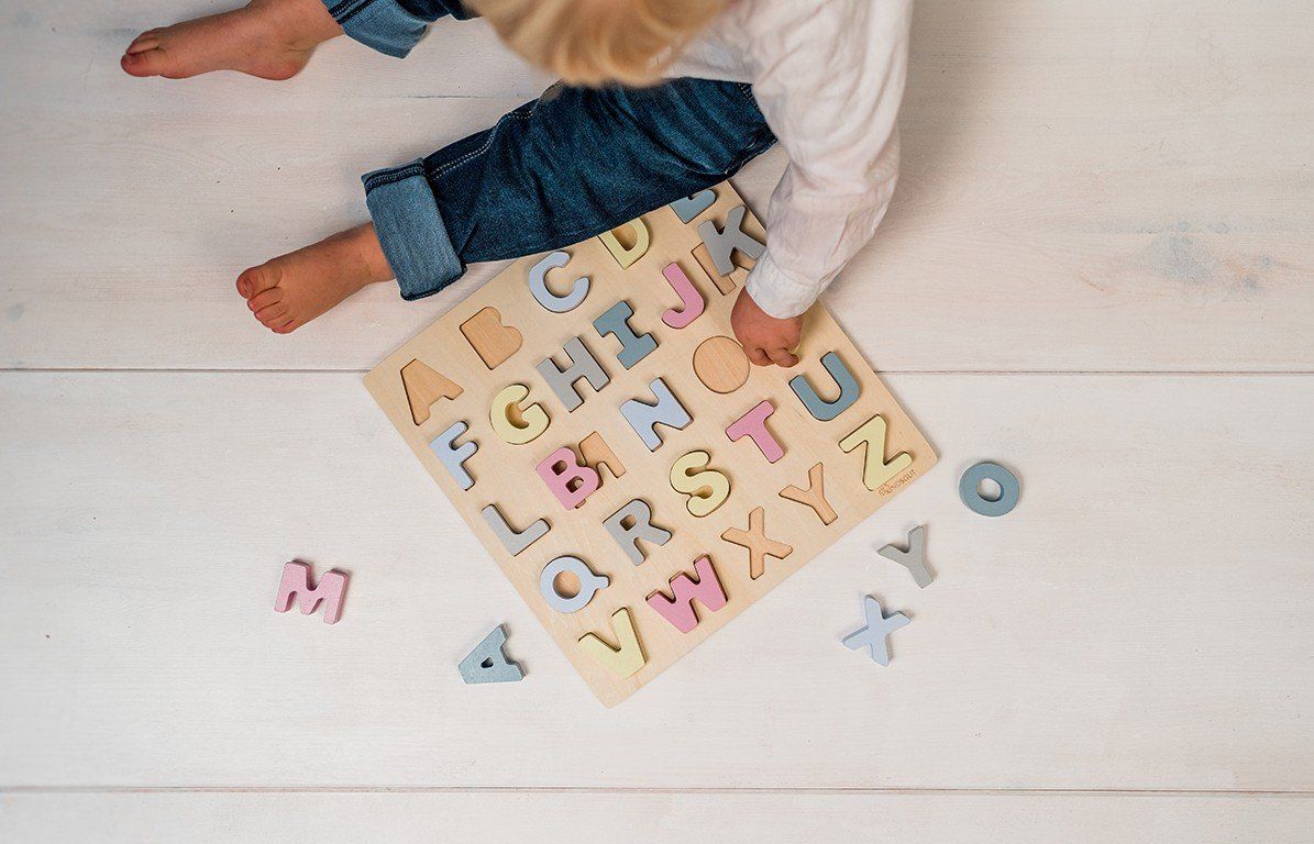 Kind sitzt neben Holzpuzzle mit Buchstaben. Einige Buchstaben sind herausgenommen. Helle Holzoberfläche.