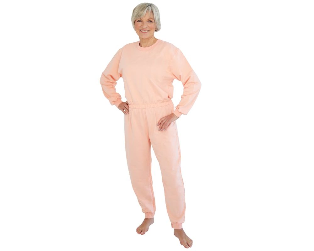 Aprikotfarbener Pflegeoverall für Frauen. Langärmliges Oberteil und lange Hose. Frau steht mit Händen auf den Hüften.