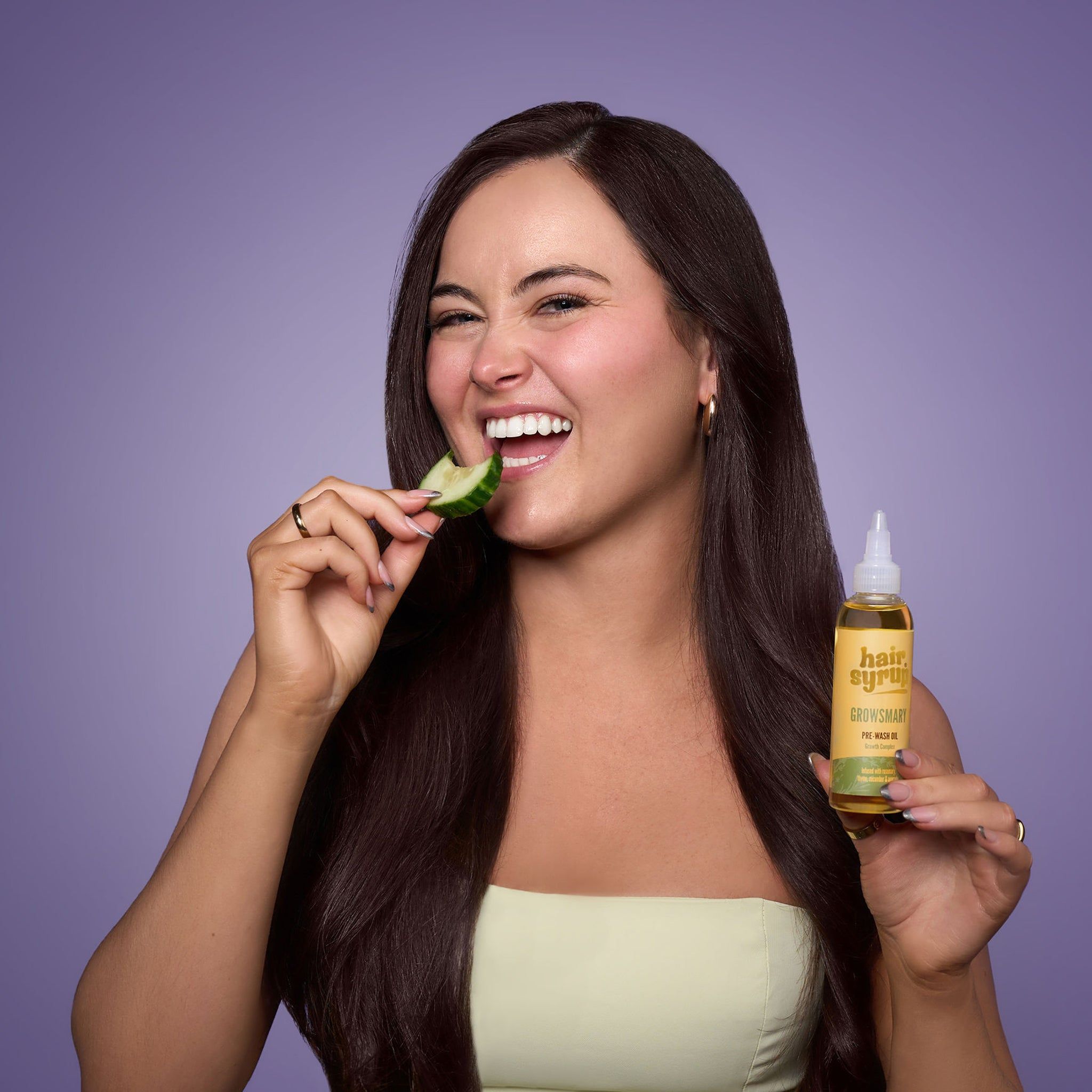 Frau mit langen, dunklen Haaren hält Flasche „Hair Syrup Growsmary“ und isst Gurke. Auf lila Hintergrund.